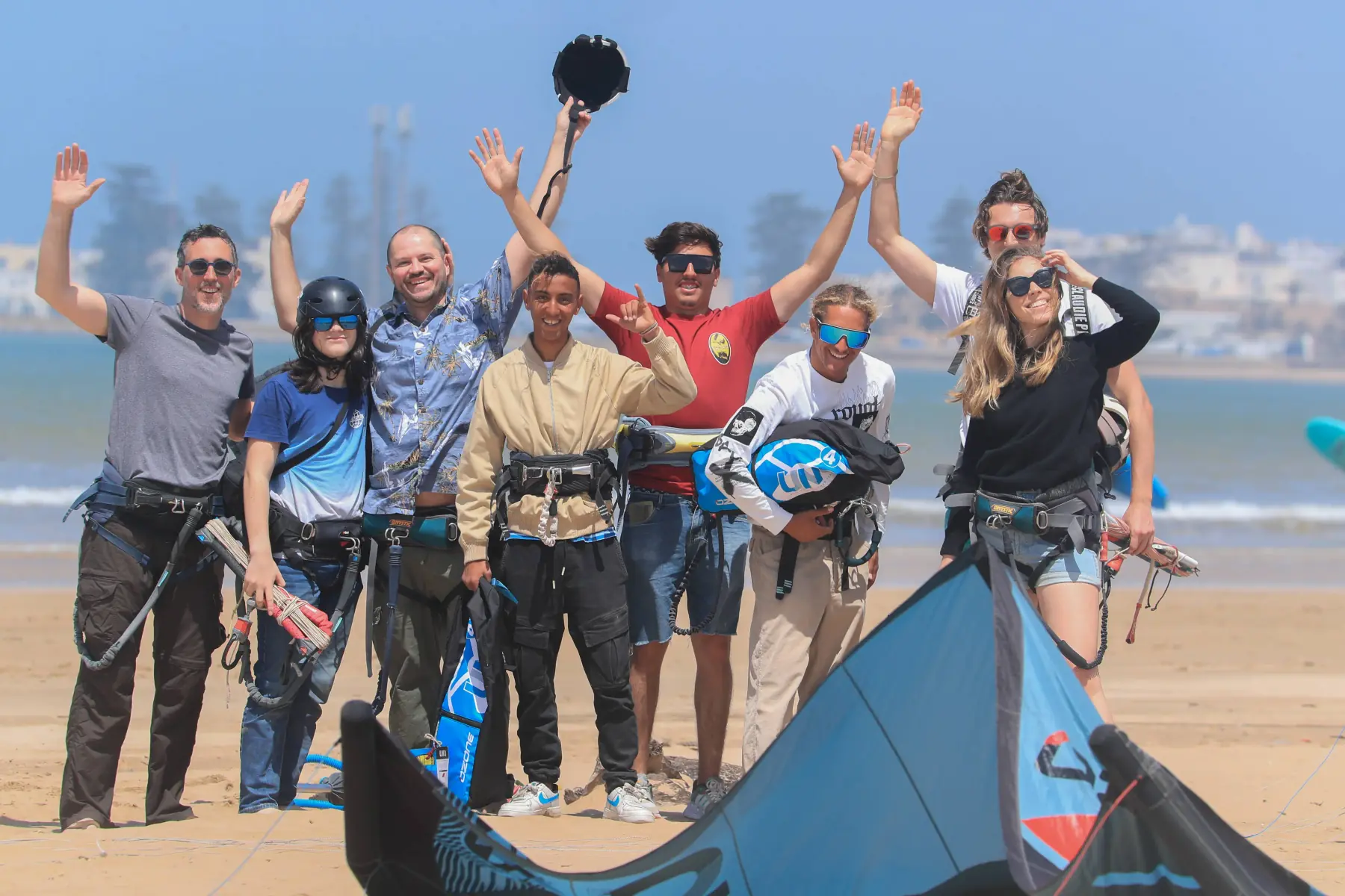 A group of people, several wearing harnesses, raise their arms and smile on the beach, with a kite lying on the sand in front of them.