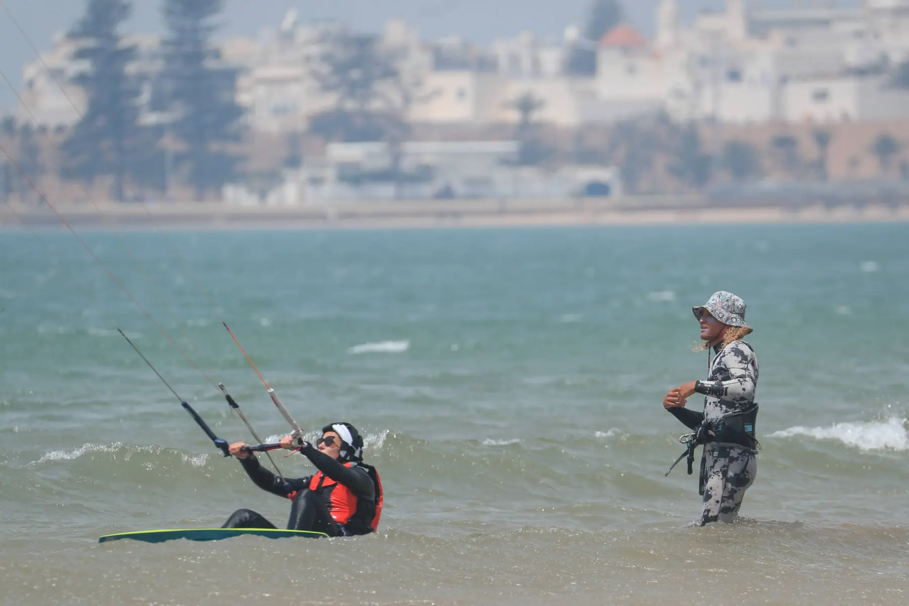 A kitesurf student sitting in shallow water with a board, holding the control bar, while an instructor in a bucket hat stands nearby. White buildings and trees are in the background.