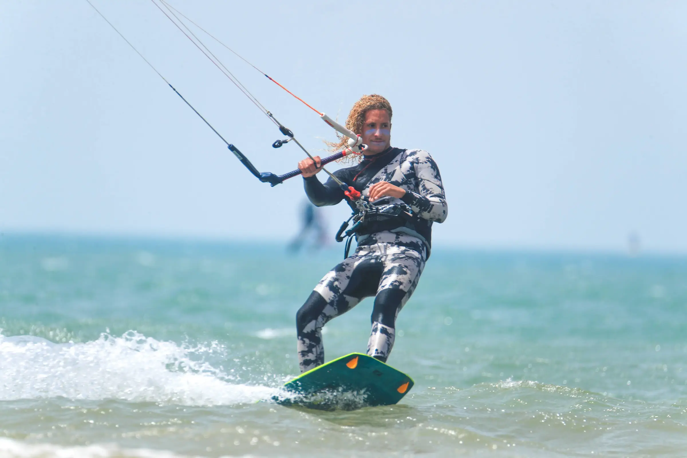 A kitesurfer in a grey and white patterned wetsuit riding a green board across turquoise water, holding the control bar.
